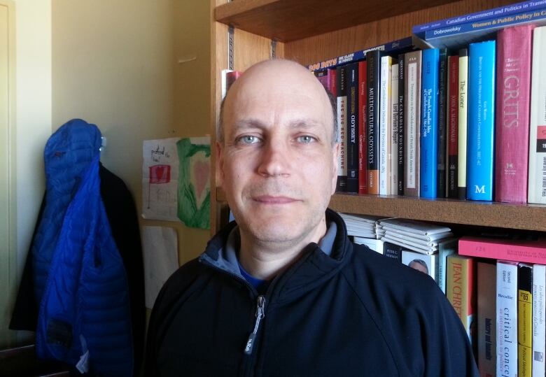 A bald man in a sweater stands in front of a full bookcase.