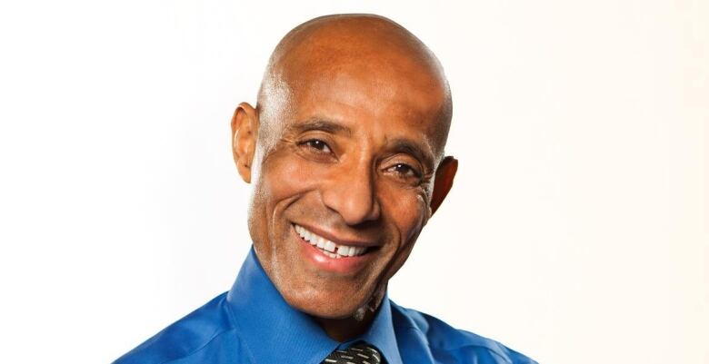 A bald Black man smiles with his arms crossed in a blue dress shirt.