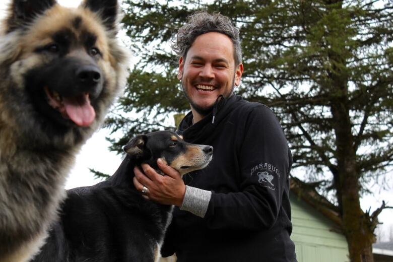 A man holds a dogs head with a smile on his face in front of trees.