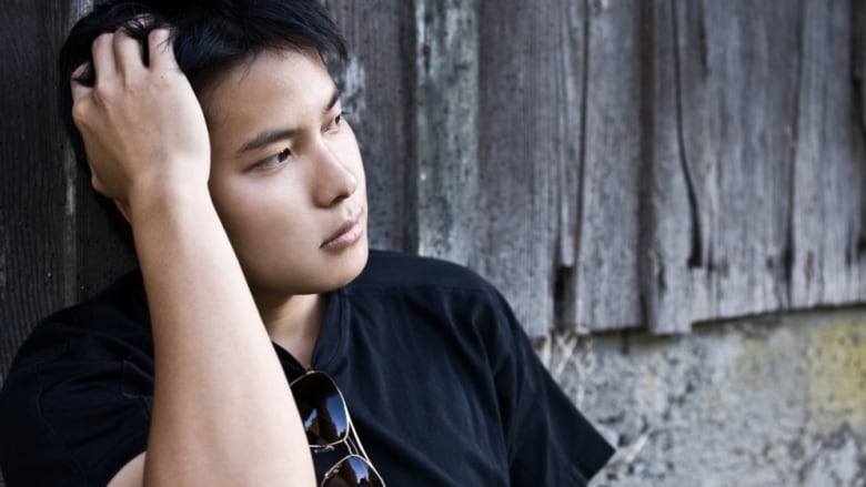 A young man sits on the ground leaning against a building, his head resting in his hand.