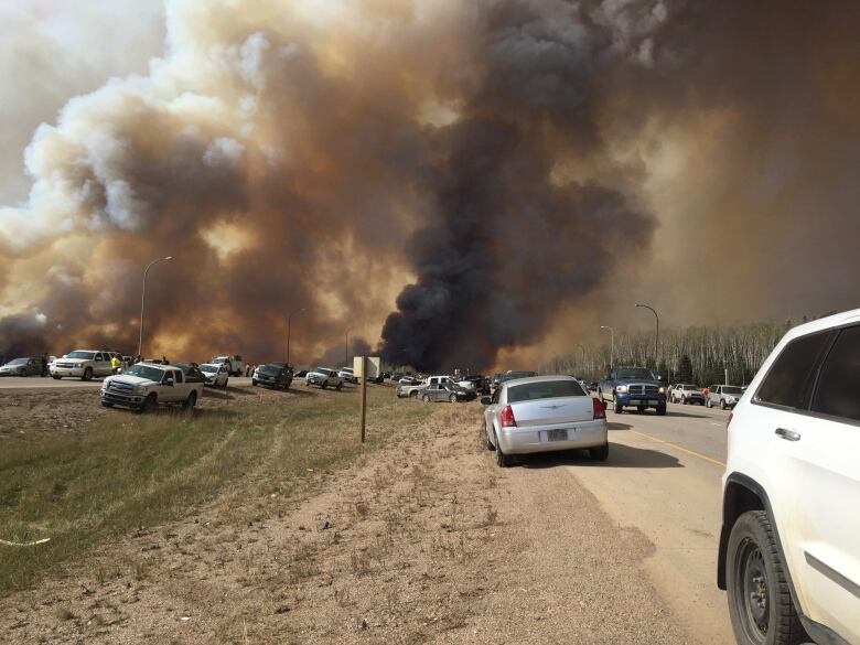 Cars are stopped on the highway and haphazardly on the shoulder and median as large black clouds from a wildfire billow overhead.