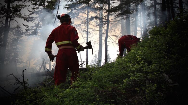 A firefighter wearing a red B.C. Wildfire Service can be seen from behind in silhouette, holding an ax, in a forest shrouded by smoke.