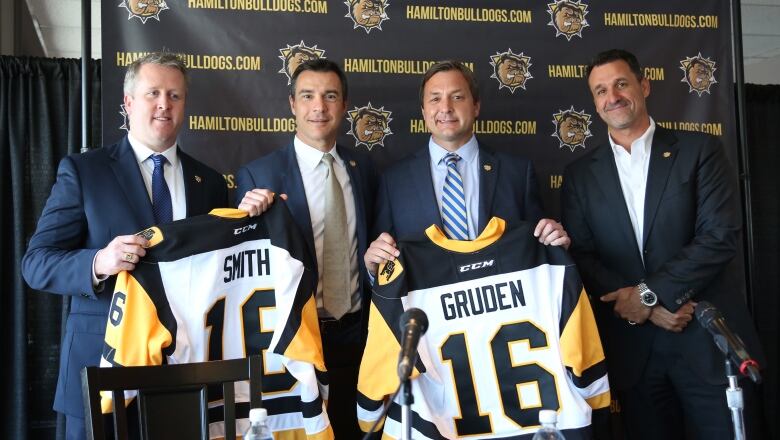 Four hockey executives and coaches at a news conference. Two are holding up team sweaters.