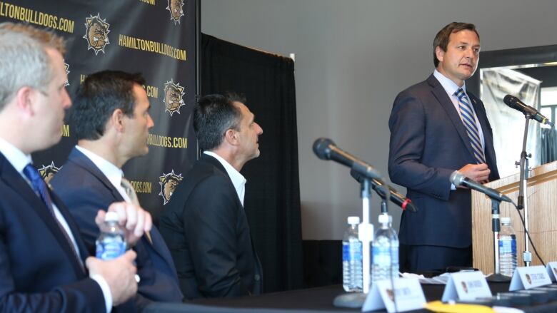 Four sports team coaches and executives give a news conference. One is speaking and three are sitting.