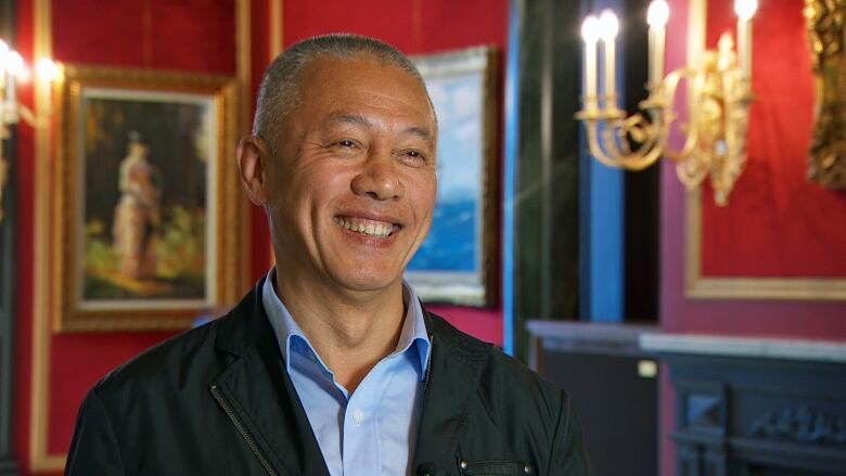 Mingfei Zhao smiles in an opulent room with paintings and wall hangings behind him.