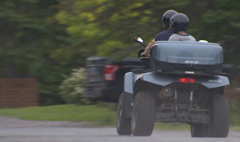 An off-road vehicle is shown from behind, moving, with two people on it, one behind the other.