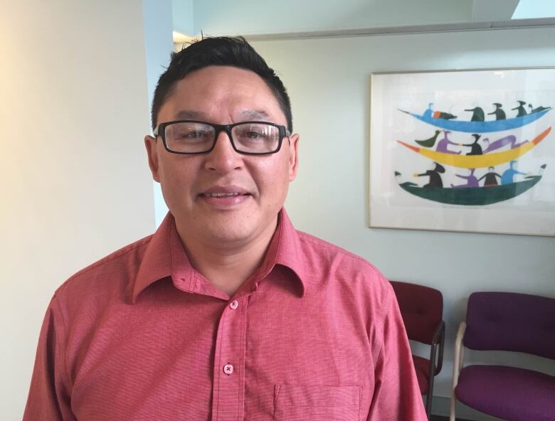 A man in a salmon-coloured shirt looking into the camera. He's wearing glasses and standing in an office room.