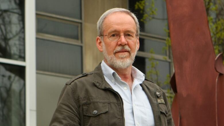 A man with a white beard and glasses stands outside a building.