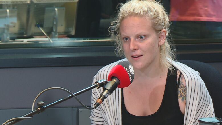 The photo of Mandi Gray was taken in 2016. She is in a CBC News studio with a radio mic in front of her. She's wearing a black tank top and stripped shirt.