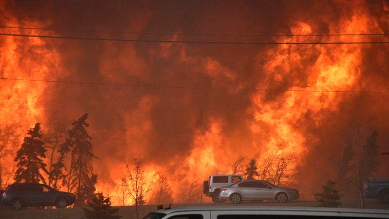 A large wild fire is blazing behind two trucks on the side of the road.