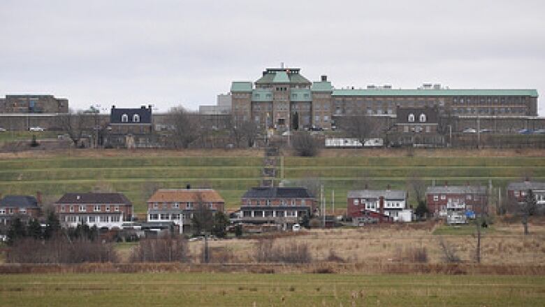 A stone prison building with a green roof is seen on grassy grounds surrounded by smaller buildings under an overcast sky.