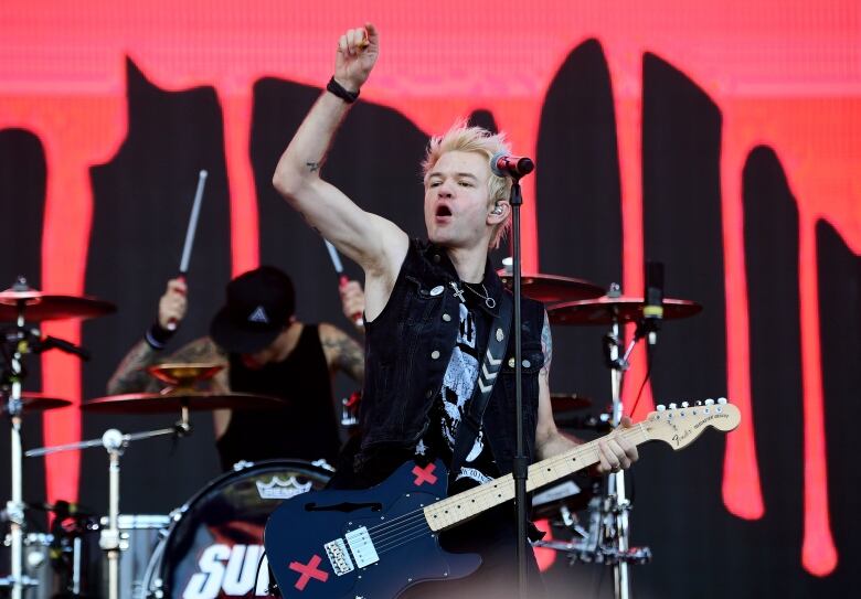 A white man dressed in black performs onstage, throwing his right arm up in the air while playing guitar.