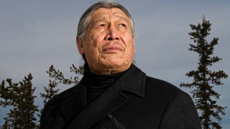 Man in black coat stands in front of trees, stares into the night.