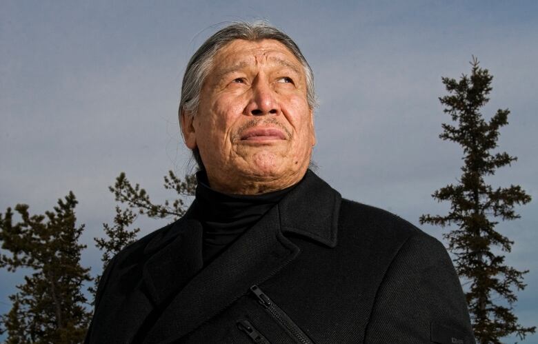 Man in black coat stands in front of trees, stares into the night.