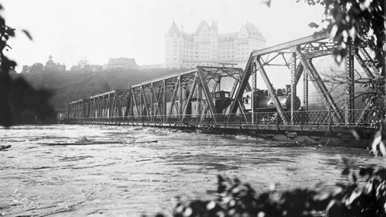 Black-and-white photo of a railway bridge with fast-flowing water right beneath it. 