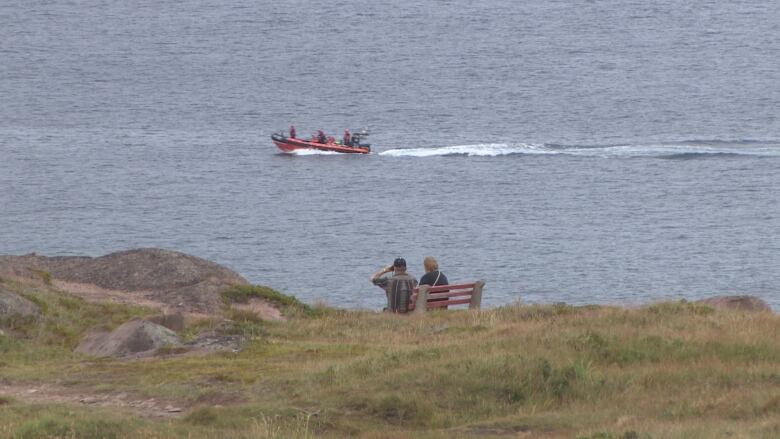 A small boat makes its way along the water. A handful of onlookers on shore watch.