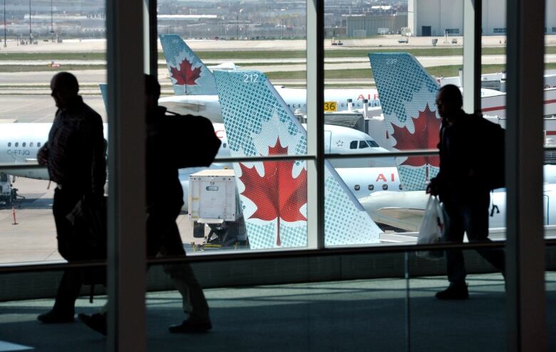 Passengers walk in an airport in front of a window. Outside, there are planes.