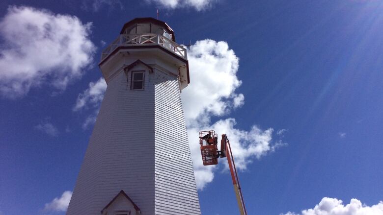 A person in a boom truck is painting a lighthouse. 