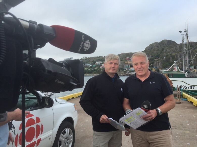 Two men in black shirts holding clipboards stand in front of a television camera.