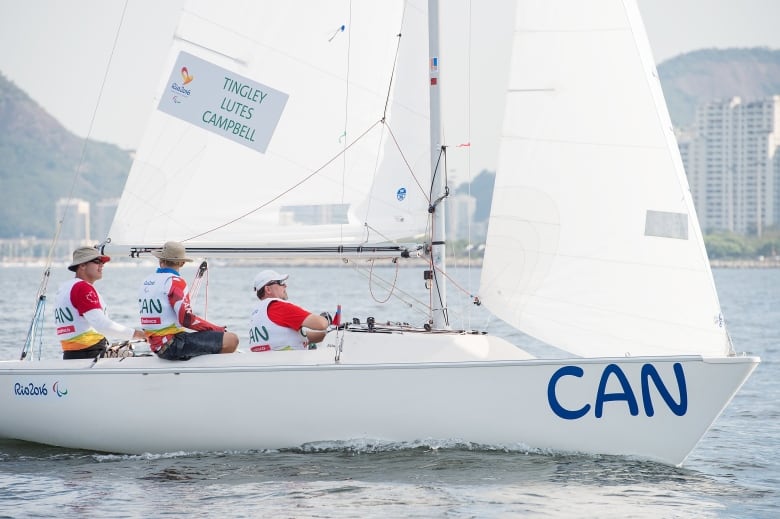 Three men sailing for Team Canada are seen sitting on a white sailboat. The sails are up, and the word CAN can be seen on the right corner of the boat.