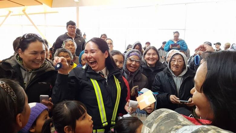 A woman smiles, holding a pin. She is surrounded by people and is holding a card. 