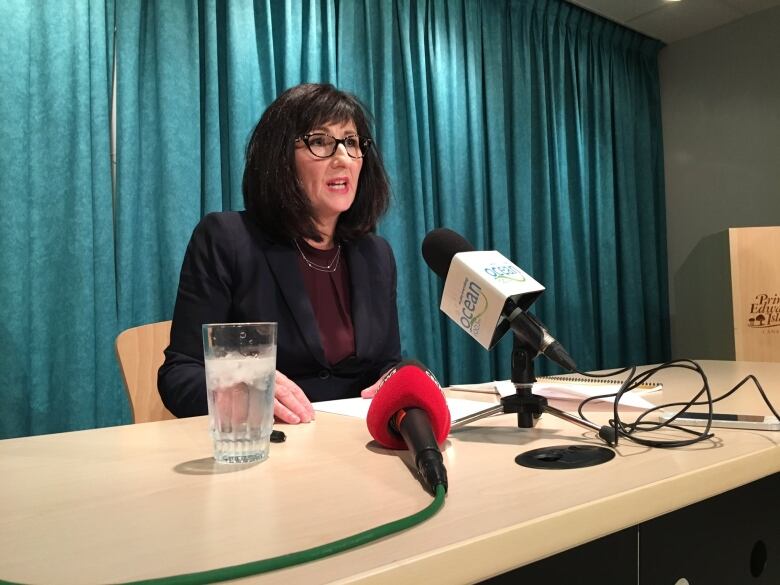 Woman with long black hair and glasses, dressed in a business suit and surrounded by microphones at a conference table.