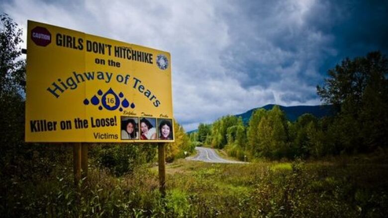 A large, yellow billboard stands beside a highway through thick forest. The billboard, marked Highway of Tears, advises girls not to hitchhike and warns of a Killer on the Loose.