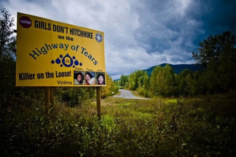 A large, yellow billboard stands beside a highway through thick forest. The billboard, marked Highway of Tears, advises girls not to hitchhike and warns of a Killer on the Loose.