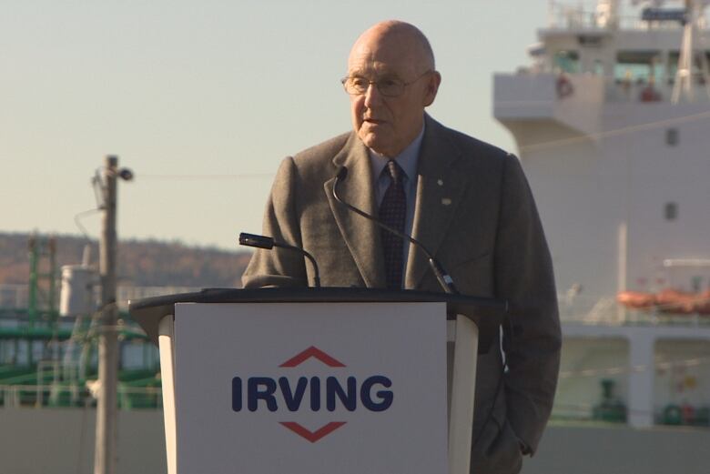 An older man wearing a business suit speaks outdoors at a lectern that features the Irving corporate logo on its front. 