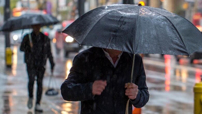 A closeup of a man walking on a city sidewalk on a rainy and windy day with his face hidden beneath an umbrella while another man holding an umbrella walks behind.