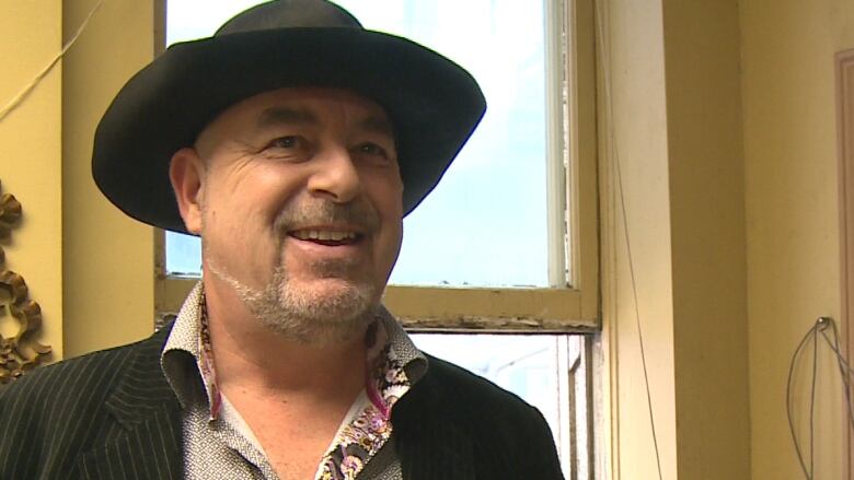 A smiling man in a hat, blazer and collared shirt.