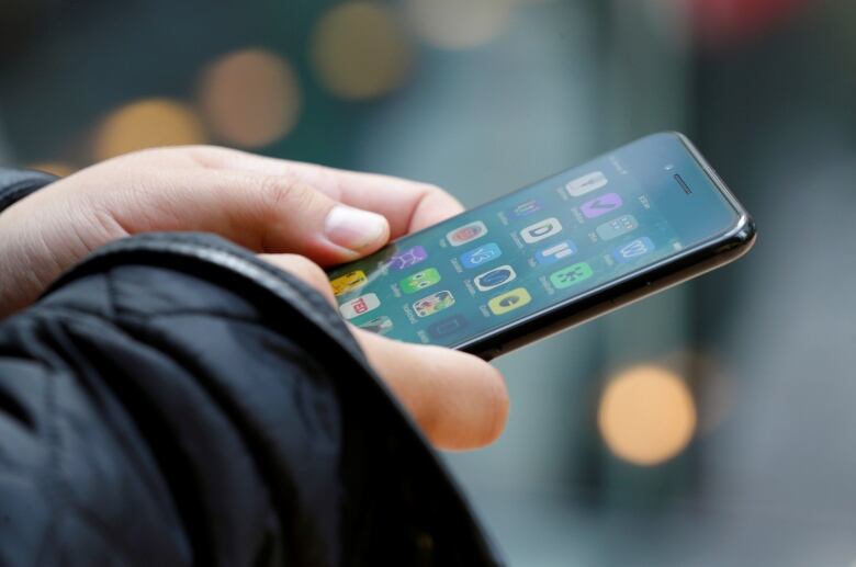 A stock photo shows a close up of two hands holding a smartphone. Apps are visible. The person is outdoors during day time.