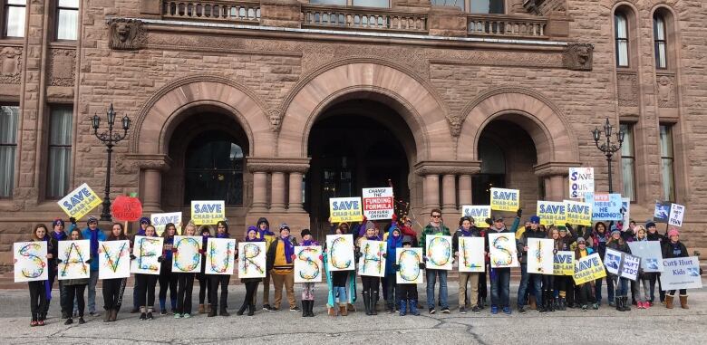 A group of demonstrators in front of the Ontario Legislature holding signs that read 