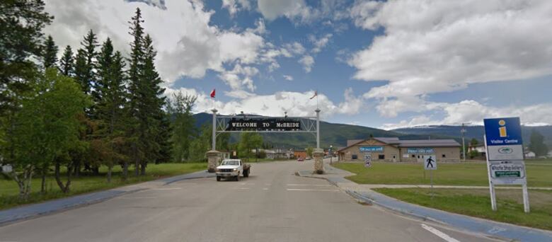 A white pick up truck drives along a highway underneath a sign that arches over the road ans says welcome to McBride.