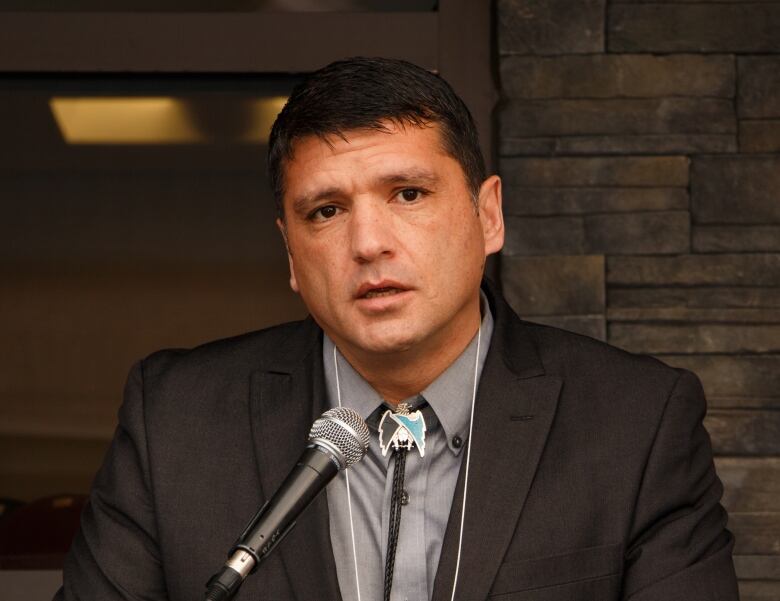 Man with dark brown hair and brown eyes wearing dark grey suit jacket and grey dress shirt sits in at table front of brick wall.