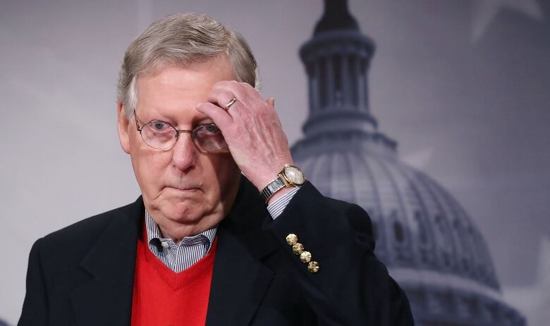 An older man in a suit gestures while standing in front of an image of the U.S. Capitol dome. 