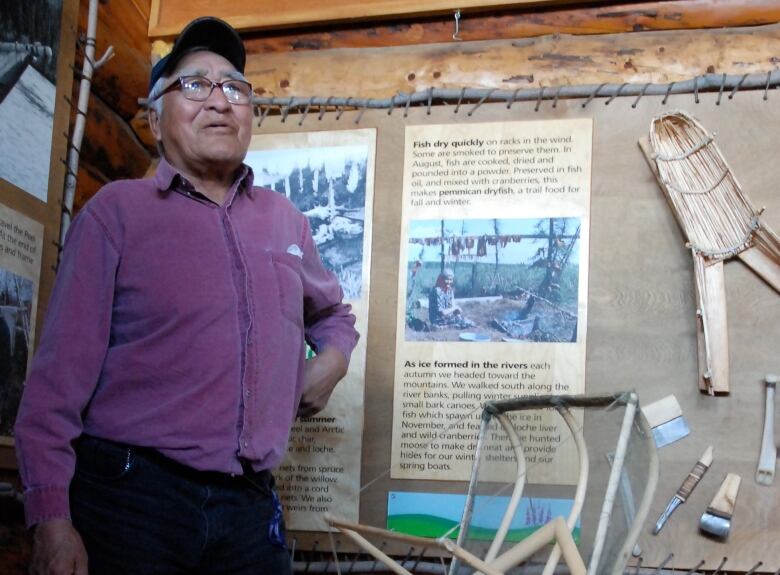 An elder stands in front of a moose hide display in a wooden cabin.