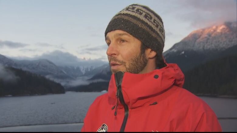 A man in red jacket and a beanie interviewing for camera with snow-capped mountain range and river on the background. 