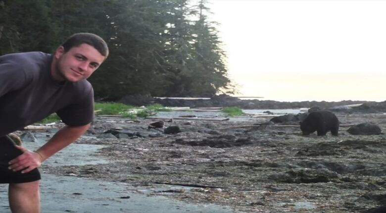 A young man, the hint of a smile on his face, leans into the frame at left on a rocky shore while a bear forages in the background. 