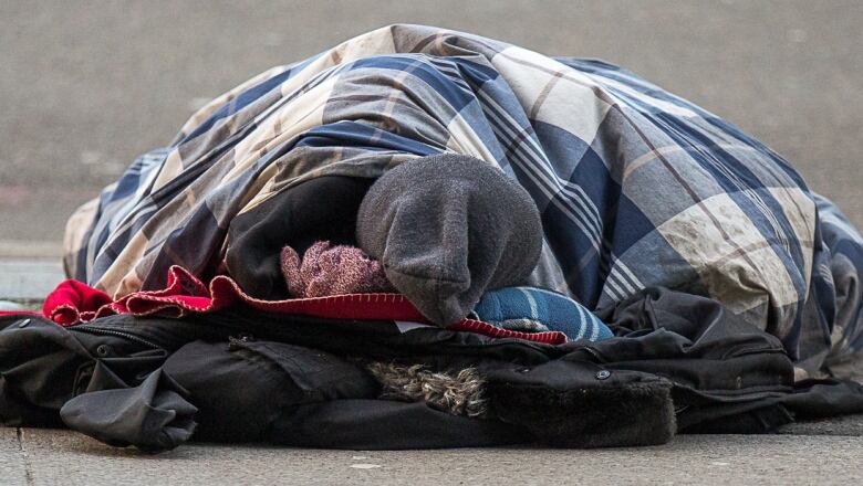 A homeless man sleeps in a sleeping bag on the sidewalk.