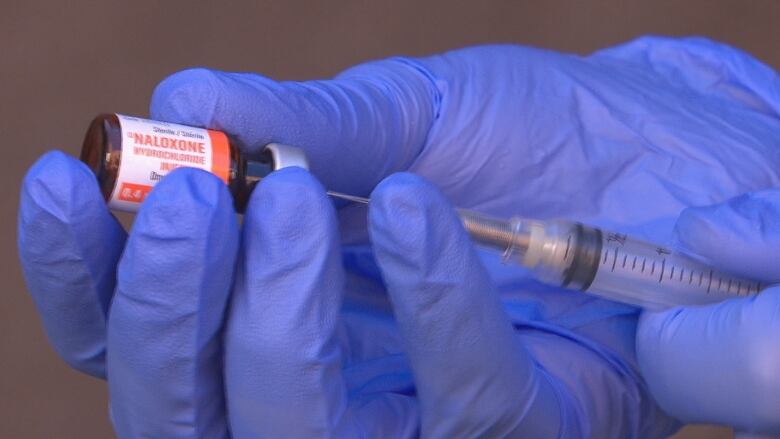 A closeup shot of two hands in purple medical gloves holding a syringe that says naloxone.