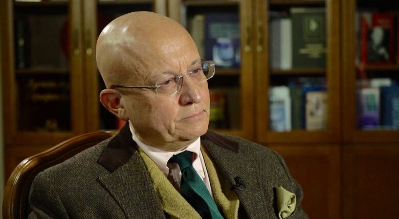 A bald man with glasses and wearing a green jacket and tie sits in front of a glass display case.