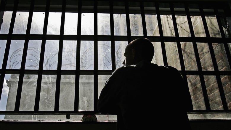 A man looks out a dirty window. Barbed wire is outside the window. 