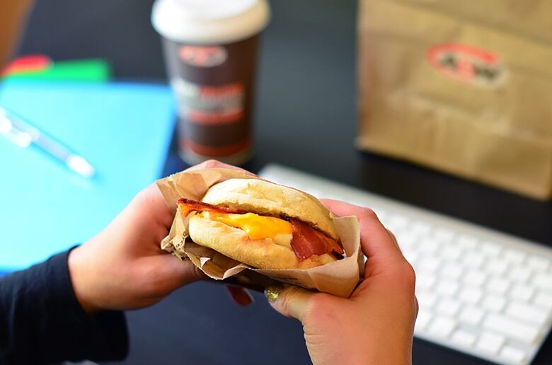 A pair of hands holds a breakfast sandwich with an A&W cup and bag and a computer keyboard in the background.