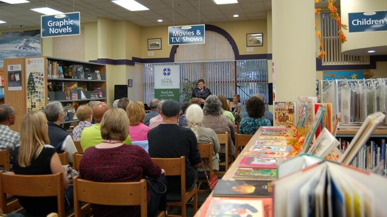 A small crowd, facing away from the camera, seated in chairs, listen to a speaker at a public library.