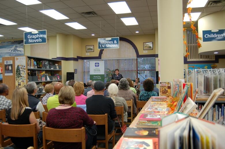 A small crowd, facing away from the camera, seated in chairs, listen to a speaker at a public library.