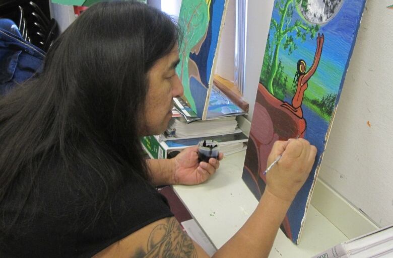 A man with long dark hair is seen painting on a canvas.