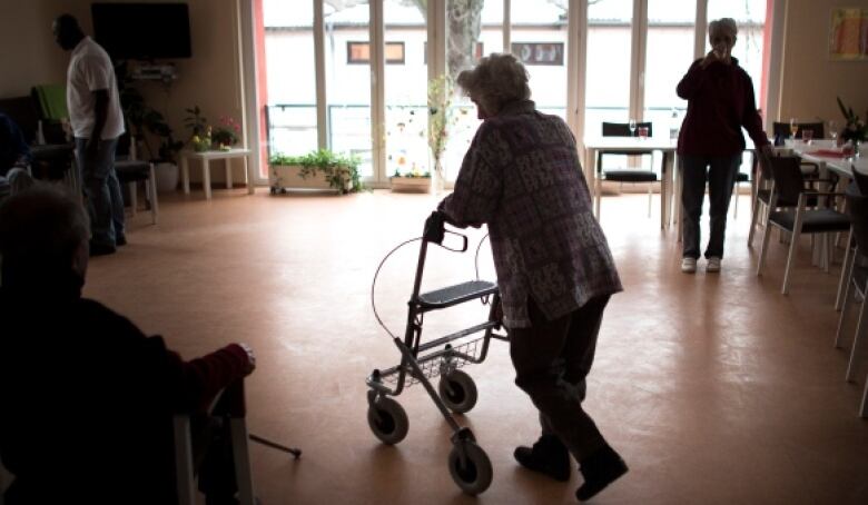 An older woman uses a walker in a room with a large window, dining tables to the right and chairs to the left.