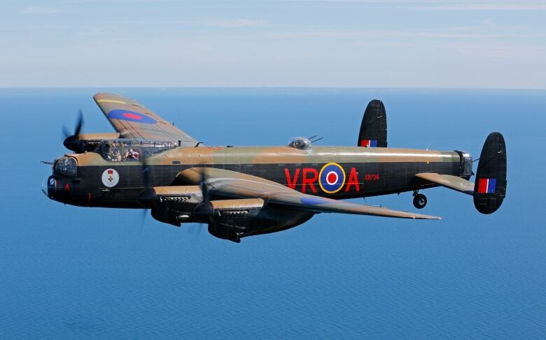 A large airplane painted in green and brown camouflage, flies over an ocean