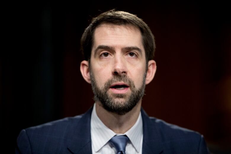 A closeup of a man with dark brown hair and a beard, wearing a suit, while speaking mid-sentence.